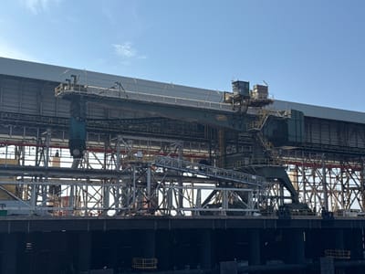 This image depicts an industrial facility, likely a port or processing plant, situated in Mersat El Hadjadj, Algeria. The primary subject is a large, dark green crane situated on a raised platform, extending over a complex network of metal structures and conveyor belts. The crane features a cab with windows and a long boom. The background consists of a vast, light-colored industrial building with a corrugated metal facade and a clear blue sky above. The foreground shows a dark, multi-tiered structure that appears to be a pier or loading area, with water visible below. The scene is devoid of human presence, suggesting the area is either unmanned or the activity is not captured in this frame. The overall impression is one of heavy industry and large-scale operations.