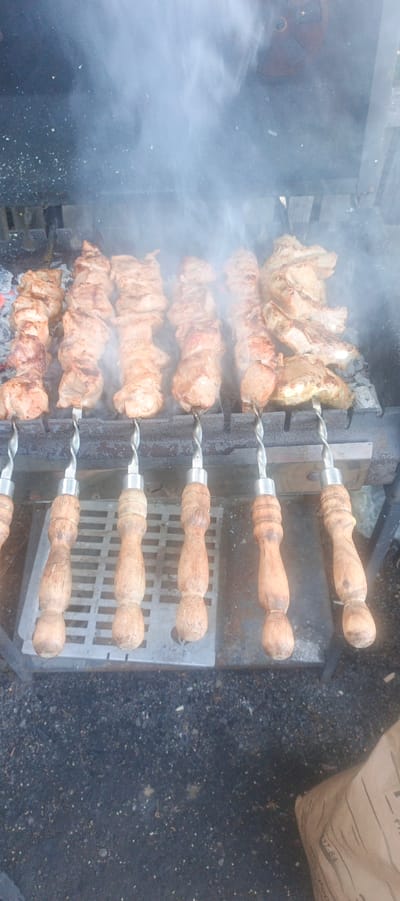 The image captures a close-up, eye-level view of several skewers of raw meat being grilled over coals. The meat, appearing to be pork or chicken, is cut into chunks and threaded onto metal skewers. Smoke billows around the skewers, obscuring some of the details of the meat and the grill itself. The skewers have wooden handles that are somewhat worn. Below the grill, a metal grate is visible, and beyond that, dark, pebbly ground. The overall lighting suggests it is daytime, possibly overcast, given the diffused light and the presence of smoke. The scene evokes a sense of outdoor cooking, perhaps a picnic or a backyard barbecue. No people are visible in the frame. There is no visible text in the image. The location context of Petukhovo, Russia, suggests a rural or suburban outdoor setting.