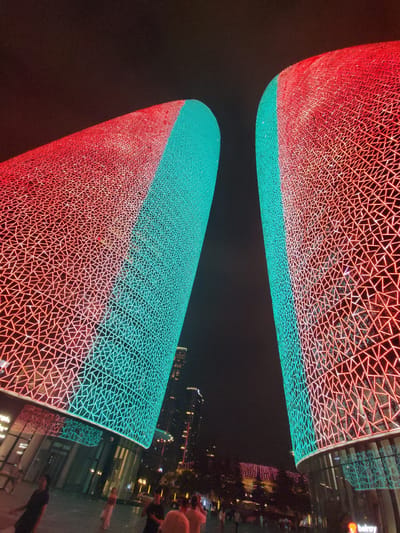 Two towering, curved structures, illuminated by red and teal LED lights forming intricate geometric patterns, dominate the night sky in Wuhou District, China. The lights transition from red on the left to teal on the right on both structures, creating a gradient effect. The textured surfaces of the buildings are composed of overlapping triangular and linear elements. Below the illuminated structures, a ground-level area with a reflective, polished surface is visible, with several individuals walking and standing. Some of these individuals are in focus, while others are blurred, suggesting movement or a shallow depth of field. In the background, other buildings with illuminated windows and lights are present, indicating an urban setting. The overall scene is dark, with the bright lights of the buildings being the primary source of illumination.