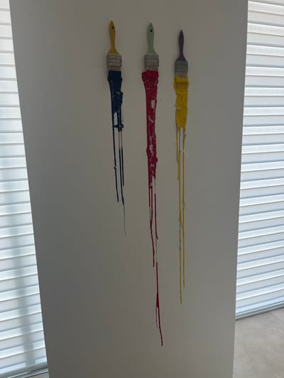 The image displays an indoor scene featuring three paintbrushes mounted vertically on a light-colored wall. From left to right, the paintbrushes are adorned with dried, dripping paint in dark blue, red, and yellow. The dark blue paint on the left brush exhibits a cracked texture with multiple thin drips extending downwards. The central brush features red paint with significant cracking and peeling along its extensive drips. The rightmost brush displays yellow paint with a smoother, yet dried and dripping, appearance. The handles of the brushes are distinct: the leftmost is yellow, the middle is light green, and the rightmost is light purple. Horizontal window blinds are visible along the left edge of the frame and partially along the right edge, indicating windows or openings providing light. Light-colored floor tiles are present in the bottom right corner. No individuals are depicted. The specific location within Galatina, Italy, cannot be determined from the image provided.