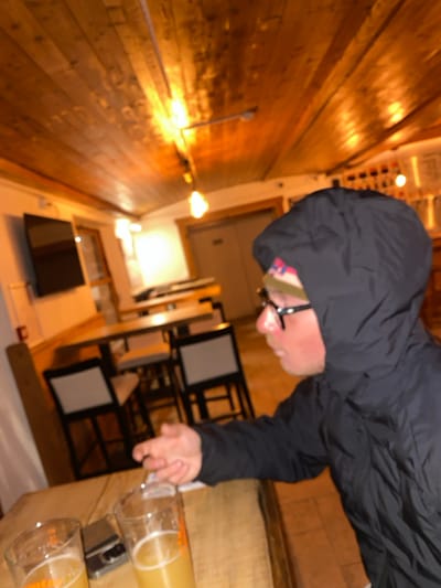 The scene depicts the interior of a commercial establishment, possibly a bar or restaurant, in Briançon, France. No specific identifying markers for the exact venue are visible. The image is blurry with warm, artificial lighting.

On the right side of the frame, a person is seated at a light-colored wooden table with a rustic edge. This individual wears a dark, possibly black, hooded jacket with the hood up, obscuring most of their hair. A multicolored beanie with pink patterning is visible beneath the hood. They wear dark-framed glasses, and their right cheek appears flushed. Their right arm is extended, with the hand reaching towards or gesturing near items on the table.

On the wooden table, two clear glasses containing a light amber or pale yellow liquid are present, consistent with beer. A dark, rectangular object, possibly a smartphone or wallet, lies on the table between the two glasses.

The background shows additional light-colored high-top tables paired with stools featuring dark frames and light-colored square seats. A flat-screen television is mounted on a white wall in the upper-left quadrant. Multiple bare Edison-style light bulbs provide warm, orange-toned illumination, one hanging centrally, others part of a track lighting system. The ceiling is constructed from horizontal wooden planks. The floor appears to be tiled. A light-colored doorway is visible in the background, along with another, darker entrance or structure.