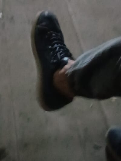 The image displays a close-up view of a person's lower leg and foot, seen from a slightly elevated perspective looking downwards. The primary focus is on a black lace-up shoe or boot worn on the left foot. The shoe features black laces tied at the top, and its sole appears to be a contrasting lighter color, possibly tan or gum brown. The person is wearing dark-colored trousers, which could be jeans or another sturdy fabric, appearing in a muted dark grey or possibly a very dark green hue. The fabric of the trousers shows some creases and folds.
The background consists of a dark, textured surface, likely concrete or pavement, which appears somewhat rough and uneven. There are faint lighter specks and variations in tone across this surface.
The overall lighting in the image is dim, suggesting low-light conditions, possibly nighttime or a poorly lit indoor environment. The image quality is somewhat blurry or soft, which could be due to the low light, camera focus, or motion. No specific activity beyond the presence of the foot and leg is discernible, nor are any emotions or visible text present.