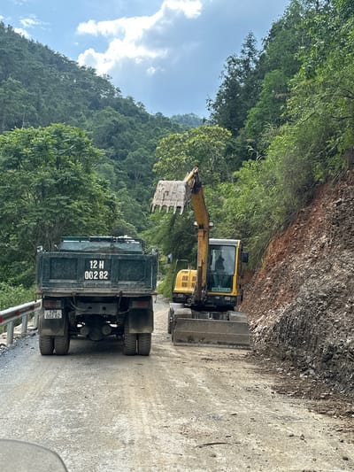 The image shows construction in the Bạch Đích Commune, Vietnam. A yellow excavator is loading dirt into the back of a green dump truck on a narrow road. The area is mountainous with dense green vegetation.