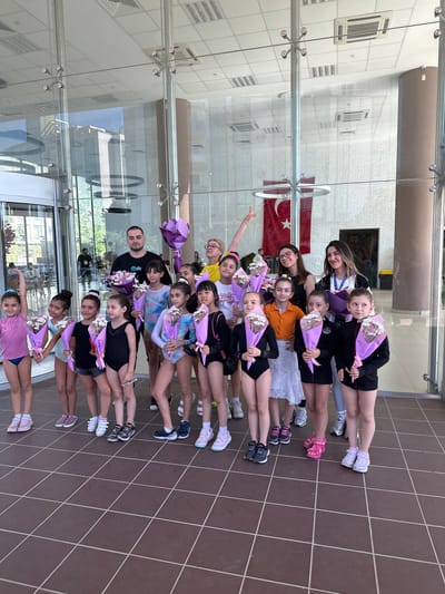 In Sancaktepe, Turkey, a group of young gymnasts, accompanied by adults, stand outside a building with a glass facade, holding bouquets of flowers. The gymnasts are dressed in various leotards, some in black, blue, and pink, with their hair styled in buns. They are posed for a photograph, holding their bouquets in front of them. Some of the adults, two men and three women, are also holding flowers. The background shows the interior of the building, with columns and glass walls, and an exterior view with greenery. A Turkish flag is visible hanging inside the building. The floor is tiled with brown square tiles.