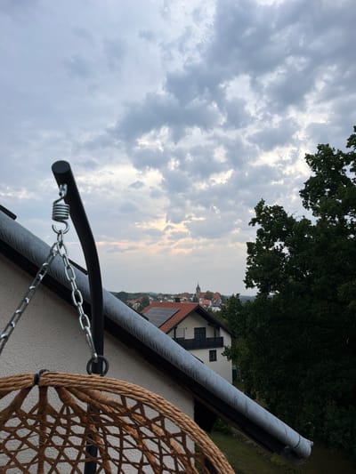 The image captures a view from a balcony or elevated structure overlooking the town of Creußen, Germany. In the foreground, the textured wicker of a hanging chair is visible, along with its supporting metal frame, spring, and chains. A grey downspout runs along the angled roofline of a white house.
Beyond the immediate foreground, a picturesque townscape unfolds under a cloudy sky. Buildings with red-tiled roofs, including a prominent church with a steeple, are clustered together. Solar panels are visible on one of the rooftops. Dense green trees frame the right side of the scene, suggesting a verdant environment surrounding the town. The overall atmosphere is subdued, likely due to the overcast weather. The image appears to have been taken during the late afternoon or early evening.