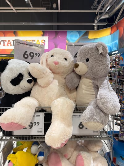 The image captures a detailed view of a retail store's toy section in Krustkalni, Latvia, specifically featuring a display of three large plush animals. In the foreground, from left to right, are a black and white panda, a cream-colored bunny, and a light grey bear, all large in scale and positioned upright within a metal display cage or shelf. The panda is on the left, looking straight ahead, with the bunny situated to its right, resting its left arm across the panda's side. The bunny features floppy ears with pink inner lining and a pink stitched nose. To the bunny's right is the grey bear, with a brown snout and a cream-colored belly, its left arm draped over the bunny. A small white tag displaying a "CE" mark is attached to the bear's left ear.
In front of the panda and bunny, a white rectangular price sign, encased in clear plastic, displays text in Latvian: "Plīša rotaļlieta panda 136cm SS26," indicating a 136cm panda plush toy. The prominent price "69⁹⁹" is printed in large black numerals. Below this, smaller text reads "Jiangsu Runshi Light and Textil," along with a barcode and a secondary price of "69,99 €/gab." Another partial price sign is visible to the right, showing "Pegaza plīša rotallieta" (Pegasus plush toy) and a partially obscured price beginning with "6." and also "69,99 €/gab."
Below the main plush toys, on a lower shelf of the metal display, various other smaller plush toys are visible, including several in yellow and some with blue and white accents. A blue plastic bin is visible to the far right. The background features colorful wall murals with large, abstract shapes in yellow, blue, purple, and orange. Above, the store's ceiling is dark grey with visible industrial-style lighting fixtures, exposed ventilation ducts, and metal grating. Other retail shelving stocked with merchandise, including packaged "Hot Wheels" toy cars, is visible further back on the right side of the frame. Latvian signs such as "ROTAĻLIETAS" (TOYS) and "AUTOMAŠĪNAS" (CARS) are discernible on the distant shelving units. No individuals are present in the image.