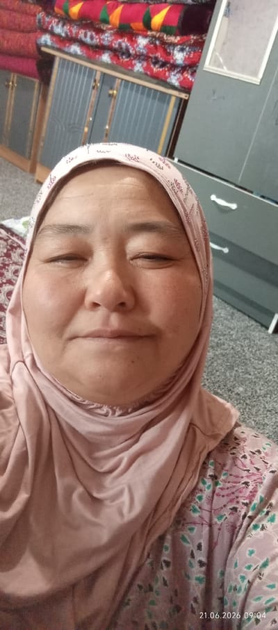 A selfie of an elderly woman wearing a pink hijab and a pink patterned dress is shown. Her eyes are closed, and she is smiling. The background appears to be a room with furniture and textiles. The date stamp on the image reads "21.06.2026 09:04". The image is believed to have been taken in Chon-Kara, Kyrgyzstan.