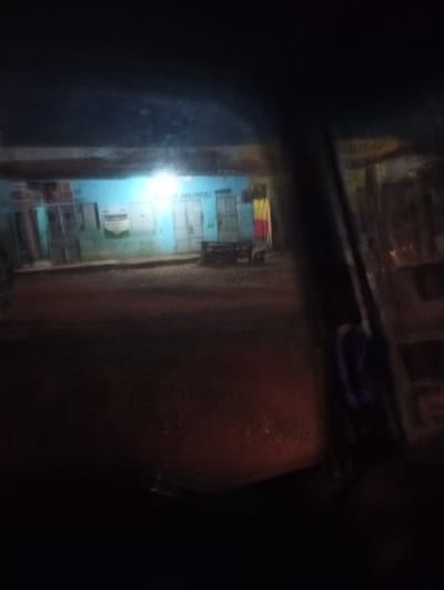 The image presents a nighttime street scene in Mururi, Kenya, viewed from inside a vehicle, likely through a windshield. The foreground is dark and blurred, with a vehicle's structural pillar visible on the right side.

In the mid-ground, a light blue-painted building stands out, brightly illuminated by a strong, central light source. This building features several dark, closed doorways or shutters. Faint white text is visible on its blue wall, appearing to read "S. WELLNESS C...", possibly indicating a business like "S. Wellness Centre". In front of the building, on the dark ground, a dark, rectangular object, resembling a bench or table, is situated with a smaller object nearby. To the right of the blue building, another structure can be seen with elements of yellow and red, which might be part of a sign or draped items.

The scene is dominated by the darkness of night, with the artificial light on the blue building being the primary illumination. The overall image has a low-light, somewhat blurry quality, consistent with photography taken from a moving or stationary vehicle at night. No people or specific activities are discernible in the static street view.