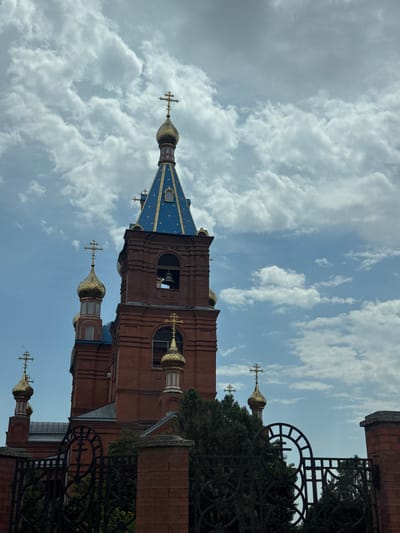 A low-angle, full-shot captures a red-brick Orthodox church in Solnechnodolsk, Russia, against a partially cloudy sky. The church features multiple golden domes topped with crosses, and one prominent blue-tiled spire with gold accents. A bell is visible in an arched opening on the bell tower. In the foreground, a decorative black metal fence with brick posts encloses the church grounds, partially obscuring lush green trees. The sky is a mix of light blue and white clouds, creating a dynamic backdrop.