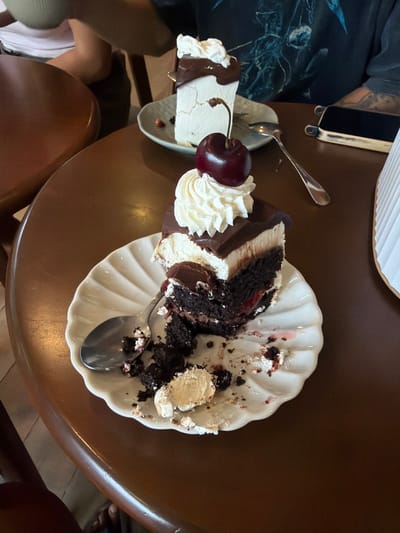 An indoor scene in Chiang Dao, Thailand, features a brown wooden table with two slices of cake and two partially visible individuals. In the foreground, a partially consumed slice of chocolate cake with a white cream layer, chocolate ganache, whipped cream, and a red cherry with a stem is centered on a white, scalloped-edge plate. A silver spoon rests within the cake's remnants, which include crumbs and red fruit stains. In the mid-ground, a second, mostly intact white cake with a chocolate topping, whipped cream, and a cherry is on a smaller light blue-grey plate, accompanied by a silver spoon. To the right of the background cake, a black smartphone with a beige case is positioned horizontally on the table. The torso and arms of a person wearing a dark t-shirt with a lighter graphic design and a tattoo on the left forearm are visible behind the smartphone. On the far left, the lower arm and knee area of another individual, wearing a light-colored top, are partially visible. A white pleated object is positioned on the far right edge of the table.