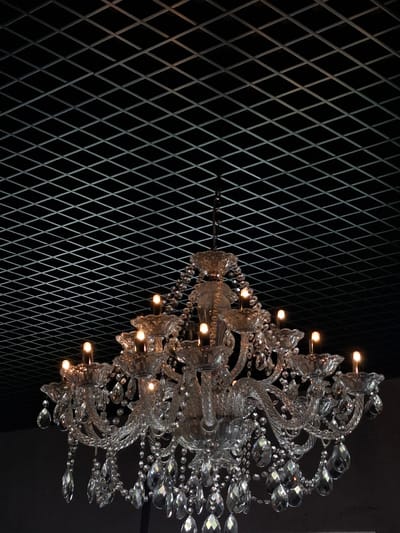 A crystal chandelier hangs from a dark, cross-hatched ceiling. The chandelier is intricate, with multiple tiers of crystal droplets and candle-shaped lights. The lights are illuminated, casting a warm glow. The background is dark, with the ceiling grid being the only visible architectural element. The image is taken from a low angle, looking up at the chandelier. The location is not discernible from the image.