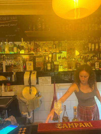 The image displays the interior of the "RECORD BAR" in Thirroul, Australia. The scene is heavily illuminated by a strong, warm yellow light emanating from a large, ridged circular fixture hanging from the ceiling on the upper right.

A female individual with dark, curly hair and a visible tattoo on her left forearm stands behind a dark bar counter, positioned towards the right side of the frame. She wears a dark tank top and a red plaid skirt. Her head is tilted downwards, and her right hand is extended towards a clear wine glass filled with ice, which rests on a red rubber bar mat embossed with "CAMPARI." Two additional bottles of liquor are visible directly behind the wine glass on the counter.

Behind the bartender, multiple shelves line the back wall, densely packed with various liquor bottles, predominantly illuminated with yellow and some green backlighting. Above these shelves, a black chalkboard displays "RECORD BAR" in white lettering, with "seasonal menu!" and a small bicycle drawing below. The wall behind the lower shelves features a row of black tiles above white tiles. To the left, a sink with a faucet, a dirty white towel, and a paper towel dispenser are visible. Inverted stemmed glasses hang from a rack beneath one of the liquor shelves.

Handwritten signs are affixed to the wall: one advertises "LIVE MUSIC 3 PM" with "$15 entry" and lists performers "LENA DOUGLAS & SARA KIRBY ...EBS...", while another sign mentions "35 MM FILM." A partially obscured sign above the sink reads "STRICTLY NO SELF..." The scene captures an active bar setting.