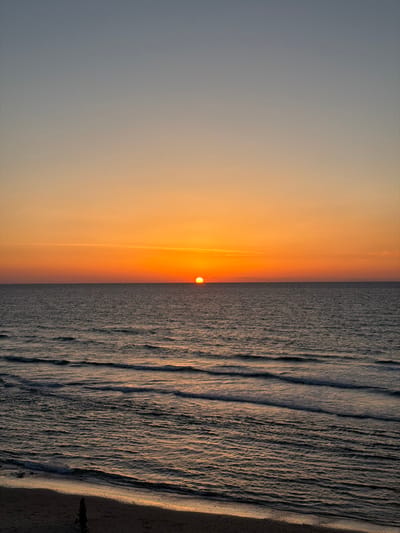 The image displays a sunset over the Mediterranean Sea, viewed from the coast of Beit Yanai, Israel. The sky occupies the upper two-thirds of the frame, showing a gradient from a light blue-grey at the top to vibrant orange and yellow hues at the horizon. A distinct, circular, orange sun is positioned centrally on the horizon line, partially set into the sea. The water of the Mediterranean Sea is dark blue-grey, characterized by multiple parallel wave patterns extending towards the foreground. White foam is visible along the crests of the waves closest to the sandy shore. A narrow strip of dark sand, reflecting subtle orange light from the setting sun, is visible at the bottom of the image. A dark, elongated, vertical object, possibly driftwood, is partially visible on this sand strip in the lower-left corner. No human figures or discernible activities are present.
