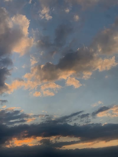 The image displays a vertical composition of the sky over Bratkivtsi, Ukraine, at an apparent sunrise or sunset. The frame is entirely occupied by clouds and atmospheric light. The upper and mid-sections feature a lighter blue sky interspersed with various cloud formations, predominantly cumulus-type, exhibiting tones of white, light grey, and subtle peach or orange from ambient light. The lower portion of the sky is dominated by darker, denser stratiform clouds, primarily in shades of deep grey and blue-grey. A distinct, horizontal band of bright orange and yellow light is visible through a break in these lower, darker clouds, indicating the position of the sun just below the horizon. No terrestrial features, structures, or individuals are present within the captured scene.