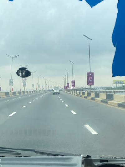 The image captures an elevated multi-lane roadway or bridge in Ebute-Metta, Nigeria, viewed from inside a vehicle looking forward through its windshield. The sky is uniformly overcast with light grey clouds. The asphalt roadway features at least two visible lanes of travel marked with white dashed lines. Concrete crash barriers, painted in alternating black and light beige rectangular blocks, delineate the sides of the road.
Numerous tall streetlights, each equipped with a solar panel, are uniformly spaced along both sides of the roadway, receding into the distance. Several of these poles carry vertical advertising banners, one prominently displaying "WEMA BANK" on the right side. Ahead, in the same direction of travel, a white van or mini-bus is visible in the distance, followed by several other indistinct vehicle silhouettes, indicating light traffic. To the right, beyond the barrier, a body of water is discernible. The distant horizon appears hazy, with faint, indistinct structures or buildings on the far left.
In the immediate foreground, the top of a vehicle's dashboard and the base of its windshield wipers are visible. An irregular, dark, amorphous spot is present on the windshield, positioned slightly above the center-left. The top right corner of the image contains an irregular blue shape, consistent with a reflection or part of the vehicle's interior frame. No individuals are explicitly depicted.