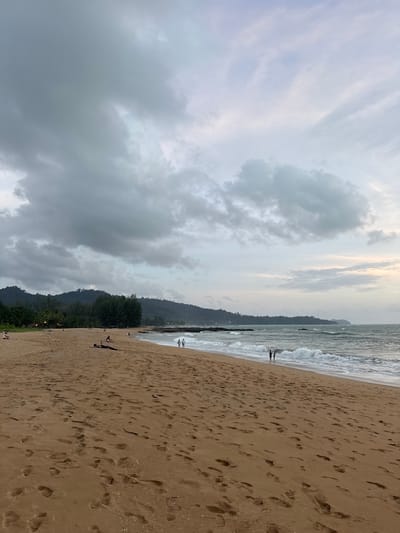 The image depicts a beach scene in Ban Bang Niang, Thailand. The sand is a warm, golden-brown color and is covered with numerous footprints. The ocean is a moderate blue-green with white waves crashing onto the shore. In the distance, a line of green, tree-covered hills forms the background. The sky is overcast with large, gray clouds.
Several people are visible on the beach. In the foreground, a person is lying down on the sand. Further down the beach, closer to the water, two individuals stand facing the ocean. A small group of people are scattered across the sand in the middle ground, some walking and others sitting. Along the tree line, lights are visible, suggesting structures or a small settlement. The overall impression is a somewhat overcast day at a tropical beach.
