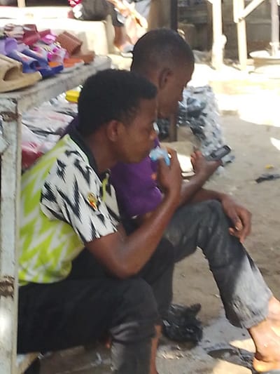 This image captures two individuals seated outdoors in Fagge D1, Nigeria. In the foreground, a person with dark skin and short dark hair wears a white, black, and lime green patterned short-sleeved shirt and dark pants, holding a light blue sachet-like object to their mouth, appearing to consume its contents. Partially obscured behind and to the right, a second person with dark skin and short dark hair wears a purple short-sleeved shirt and dark pants, barefoot, engrossed in a dark-colored mobile phone held in their left hand. Both individuals are seated on a low surface. To the left, a light-colored structural element, possibly part of a stall or bench, frames the foreground. Behind the individuals, on a raised surface, various merchandise items are visible, including multiple pairs of sandals or slippers in colors like red, purple, and beige. The ground is a light-colored, uneven surface, likely dirt or concrete, and the scene is illuminated by strong daylight.