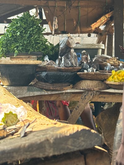 This image depicts a bustling market scene in Kagwaru, Nigeria. The foreground is dominated by a rough wooden surface with some scattered green and purple items, possibly leaves or flowers. Behind this, a stall displays a variety of goods. A large pile of fresh green leafy vegetables is prominently featured in a dark basin. Next to the vegetables, several woven baskets hold assorted items, some wrapped in plastic bags. One basket contains what appears to be dried fish or other food items. In the background, more baskets, some filled with goods and others empty, are visible, along with what looks like a large plastic container or tub. Partially visible in the background are the legs of individuals, one wearing a red and white patterned garment, suggesting active commerce. The overall environment appears to be an outdoor market with a rustic, makeshift stall construction.