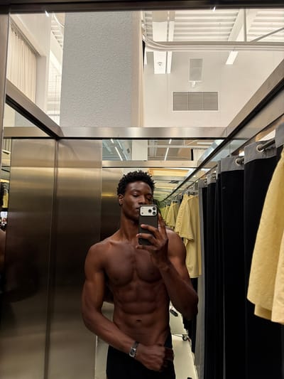 A shirtless, muscular Black man is taking a selfie in a dressing room. He is holding a phone with a triple-lens camera in his right hand, positioned to capture his upper torso and face. He is wearing a wristwatch on his left wrist and dark-colored pants. The dressing room has shiny, vertical metallic panels. Above him are clothes hanging on a rack. The reflection in the mirror shows a glimpse of what appears to be a store interior with white fixtures and geometric design elements. The location, Fibracolor, Spain, cannot be confirmed from the details of the image alone.