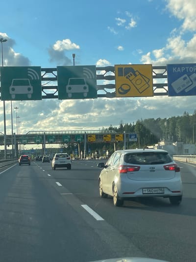 The image shows a view from inside a car driving on a highway towards a toll collection point. In the foreground, a white Kia hatchback is in the right lane. Several other cars are visible ahead, all traveling in the same direction. Overhead, a series of signs indicate toll lane options. The signs feature pictograms of cars with radio waves, suggesting electronic toll collection, and a sign with money and a credit card, indicating payment options. The sky is blue with scattered clouds, and trees are visible in the background. The location is identified as Pesochny, Russia.