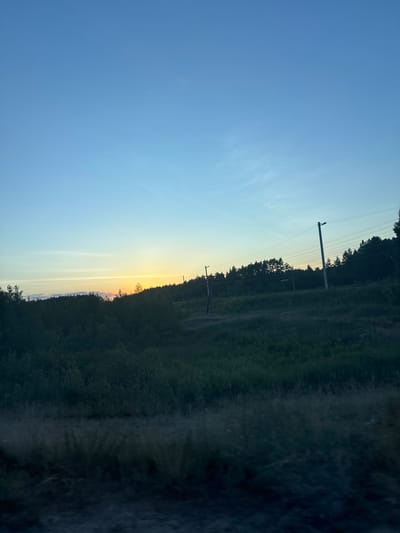 The image depicts a landscape scene in Orlivka, Ukraine, captured during either sunrise or sunset. The sky transitions from a clear, bright blue on the left to a soft yellow and orange hue near the horizon on the right, indicating the presence of the sun. A line of dark trees forms a dense border along the right side of the frame, suggesting a wooded or forested area.
Several utility poles with power lines are visible within the treeline, stretching across the frame. These poles suggest the presence of infrastructure within or near the natural landscape. The ground beneath the trees appears to be covered in vegetation, with some hints of a path or road running through it. The overall impression is one of a rural or semi-rural setting during a transitional period of the day. No people are visible in the image. The weather appears to be clear, with no clouds or precipitation. The emotions conveyed are tranquil and serene, characteristic of natural light phenomena at dawn or dusk. No text is visible in the image.