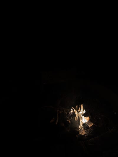 A small campfire burns brightly in the darkness, illuminating a patch of ground made of dirt and wood debris. The flames are a vibrant yellow and orange, with wisps of smoke rising upwards. The surrounding area is completely dark, creating a stark contrast with the illuminated fire. The scene is likely taking place outdoors, possibly in a rural or natural setting. The exact location within the city of Kierwik, Poland, is not determinable from the image.