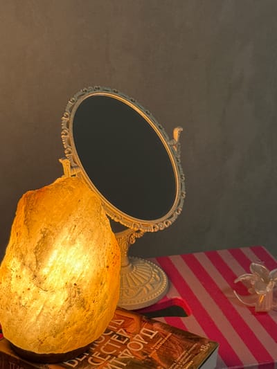 This image features an ornate oval mirror with a decorative, gilded frame. Positioned in front of the mirror is a salt lamp, casting a warm, yellow-orange glow that illuminates a textured, crystalline surface. To the right of the salt lamp, a pink and white striped gift box with a sheer ribbon flower is visible, partially obscured by a book. The book's cover displays text in Cyrillic script, with some lettering appearing in bold. The "18+" symbol is also clearly visible on the book, indicating mature content. The background is a dark, textured surface, possibly a wall. The scene appears to be indoors, with soft lighting creating a cozy and intimate atmosphere. No people are present in the image. The overall impression is one of decorative arrangement and an implied intimate or personal setting. The text on the book is partially decipherable, including "DEMOC," "CECTER," "ИТΦΟΡΩ," "В ПЕРЕВОДЕ," "АННЫ БАБЯШКИНОЙ," "ФОРД. ГЛАМУРНЫх," "ЭПЕЯ, В КОТОРОЙ СПЛЕЛИСЬ," "» И «КОРОНА»," and "MERICA."