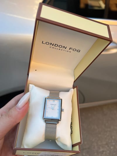 In the city of Heinsberg, Germany, a rectangular wristwatch with a silver mesh band and a pearl-white face is presented in an open, cream-colored box with brown trim. The watch face features Roman numerals, diamond-shaped hour markers, and a silver second hand and minute hand. The London Fog logo is visible on the watch face. The box is resting on a surface, possibly a car, as suggested by the reflections and the metallic sheen of the background. A person's hand with long, light-colored fingernails is holding the box, partially obscuring the lower left corner. The lighting indicates it is daytime, with natural light illuminating the scene.