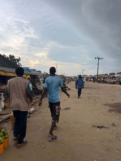The image depicts a street scene in Oko Amakom, Nigeria. The ground is unpaved and dusty, with scattered debris. Several people are visible, walking and standing. In the foreground, two men are walking away from the camera. The man on the left is wearing a patterned sleeveless vest and dark pants. The man on the right is wearing a blue t-shirt and checkered shorts. Further down the street, other individuals and market stalls can be seen. Vehicles, including what appears to be a tricycle and a van, are present in the background. Power lines and poles are visible against a cloudy sky. The overall impression is that of a bustling outdoor market in a somewhat informal urban setting.