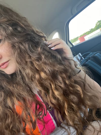 The image shows a close-up of a woman's head and shoulder, with her long, wavy, brown hair dominating the foreground. She is sitting in the passenger seat of a vehicle, as evidenced by the car interior visible in the background, including a window looking out onto a street with trees and buildings. The woman is wearing a brightly colored t-shirt with abstract designs. Her left hand is raised, touching her hair, and her fingernails are painted with a light, shimmery polish. A backpack is visible behind her. The image appears to be taken during daylight. The location is identified as Trilistnik, Bulgaria.