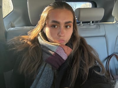 A woman with long brown hair is visible from the chest up. She wears a dark-colored sweater and a plaid scarf with shades of gray, maroon, and brown. Her face is centered in the frame, and she looks directly at the camera. The setting appears to be the back seat of a car, with gray leather seats and a headrest visible. Light illuminates the scene from the side, creating a strong contrast on her face and hair.