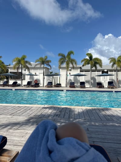 The image presents a ground-level, first-person perspective of a poolside area at a resort in San Pedro Town, Belize. In the immediate foreground, the legs of an individual, partially draped with a light blue towel, are visible over a weathered wooden deck. The deck exhibits strong shadows cast by overhead palm trees. A rectangular swimming pool with clear turquoise water and a blue tiled edge occupies the midground; numerical depth markers "3", "4", and "5" are discernible on its perimeter. Beyond the pool, a continuous row of single-story, horizontally paneled white buildings with light grey roofs forms the backdrop. In front of these structures, multiple dark grey sun umbrellas are open, shading dark lounge chairs positioned on the ground. Several mature palm trees are interspersed among the buildings and umbrellas, with their green fronds extending into the bright blue sky. An orange lifebuoy is affixed to the wall of one of the central white buildings. The sky above is largely clear, with scattered white cumulus clouds. The scene is brightly illuminated by natural sunlight.