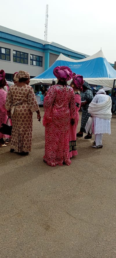 The photograph depicts a group of women in Goron Dutse, Nigeria, gathered on a paved surface. They are dressed in brightly colored, patterned traditional attire, with elaborate headwraps. In the background, there is a building with blue trim and a large white and blue tent. A communications tower is visible in the sky above the building.