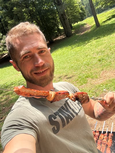 A male individual is depicted in a close-up, outdoor selfie, situated in Dolovi, Montenegro. The subject has short, spiky blonde hair and a light brown beard and mustache, smiling directly into the camera. He wears an olive green t-shirt with the upper portion of dark lettering, appearing to read "STAY...", visible on his chest. His left forearm displays a multi-colored tattoo. He holds a metal skewer horizontally with his right hand, presenting several pieces of grilled meat, consistent with shashlik or kebab, interspersed with sliced onions. The meat pieces exhibit a reddish-brown, slightly charred exterior. In the lower right quadrant of the image, a charcoal grill (mangal) is visible on the ground, supporting multiple additional metal skewers loaded with similar grilled meat and onions, with subtle indications of rising heat or smoke. The immediate surroundings consist of green grass and dried earth. The background features a sloping, grassy area with a visible dirt path ascending uphill, flanked by mature deciduous trees under bright sunlight, which casts distinct shadows. The scene is illuminated by strong natural daylight, suggesting a clear, sunny outdoor environment.