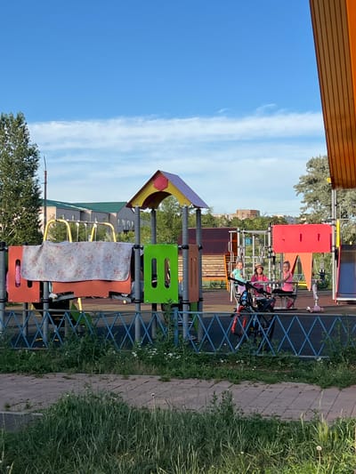 The image depicts a playground in Stepnogorsk city administration, Kazakhstan, bathed in the warm light of a late afternoon sun. The sky is a vibrant blue with wispy white clouds. A modern playground structure, painted in bright red, orange, yellow, and green, stands centrally. A patterned blanket is draped over a section of the playground equipment.
Several young girls are visible on the playground. Three girls are seated on a swing-like apparatus. One girl, wearing a pink top and a straw hat, is in the center. To her right is a girl in a pink top, and to her left is a girl in a turquoise top. A pink scooter is positioned to the right of the swing. A bicycle with red accents is parked in front of the playground structure.
The background reveals residential buildings with green roofs and other structures, suggesting an urban environment. Trees with green foliage surround the playground. The ground of the playground appears to be covered with a dark material, and a paved walkway with interlocking stones is visible in the foreground, bordered by overgrown grass and small white flowers. A blue metal fence with a diamond pattern separates the walkway from the playground. The overall scene conveys a sense of a typical summer day in a residential area.