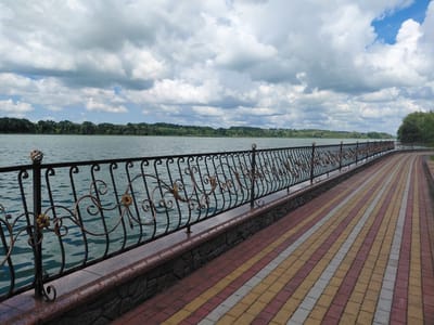 The image shows a waterfront promenade in Kovalivka, Ukraine, under a cloudy sky. A decorative black metal fence with ornate gold accents separates the promenade from a calm body of water. The promenade itself is paved with interlocking bricks in a pattern of red, yellow, and white. In the background, a treeline with lush green foliage borders the water, and the sky is filled with dramatic, textured clouds. The scene conveys a sense of public space and natural beauty.