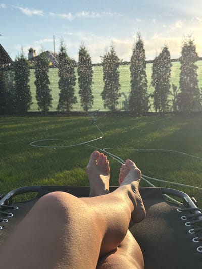 The media file shows a close-up view of a person's legs and feet resting in a black recliner. The recliner is on a green lawn, which is being watered by a sprinkler system. In the background, there are trees and a house. The sun is setting, casting a warm glow on the scene. The sky is blue with a few white clouds. The person's feet are bare, and their toenails are painted pink. The overall mood is peaceful and relaxing. The scene appears to be in a backyard in Kurylivka, Ukraine.