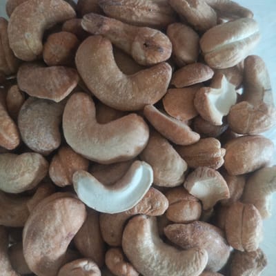 The image displays a close-up, overhead view of a pile of roasted cashew nuts. The nuts are various shades of light brown and tan, with some exhibiting a slightly darker, more roasted appearance. They are irregularly shaped, as is typical for cashews, with some showing the characteristic curved shape and others appearing more oblong. The texture appears somewhat rough and dry. The lighting is even, and the background is a light, neutral color, likely a table or surface. There are no people, actions, or interactions depicted. The image is a still life focusing solely on the food item. There is no information in the image to suggest its location in Los Teques, Venezuela.