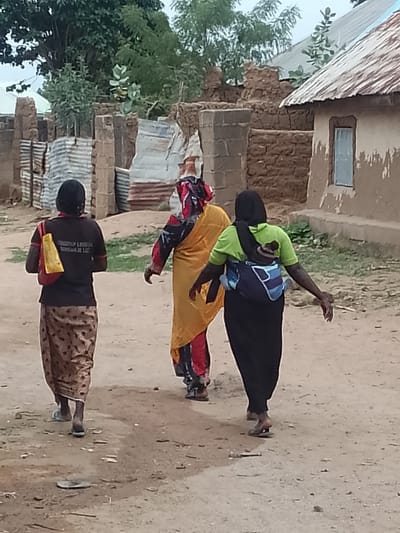 Three women are walking away from the viewer on a dirt path. The woman on the left has a black shirt, patterned skirt, and a yellow bag on her shoulder. The woman in the middle is wearing a yellow robe with red and black patterned sleeves and hood. The woman on the right is wearing a light green shirt and black skirt and has a baby strapped to her back. In the background are buildings made of mud and corrugated metal. This image was taken in the city of Biliri, Nigeria.