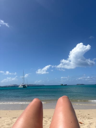 The image depicts a first-person perspective scene from a sandy beach in Spanish Town, British Virgin Islands, on a clear, sunny day. The lower half of two human legs, appearing tanned, are visible in the immediate foreground, positioned on light beige sand. Beyond the sand, gentle waves break on the shoreline, leading into a body of turquoise-to-deep-blue ocean water. In the mid-ground, a white catamaran sailboat with a single mast is anchored to the left, and a large white motor yacht is anchored further to its left. To the right of the catamaran, a dark, partially submerged or derelict vessel is visible. Multiple distant green-blue landmasses, characteristic of islands, line the horizon. The sky is bright blue with several scattered white cumulus clouds, including a large, distinct formation in the upper right quadrant. No explicit actions or interactions are depicted beyond the implied reclining position of the individual whose legs are visible.