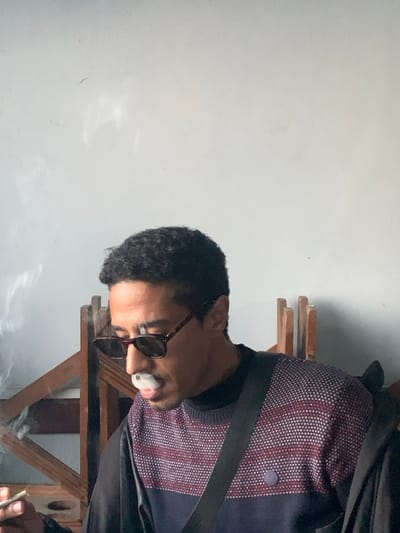 An upright, medium shot captures a man in profile, facing towards the left side of the frame, against a plain light-colored (white or light grey) wall. He has dark, short curly hair and is wearing brown-framed sunglasses. The man is dressed in a dark undershirt beneath a distinctive sweater that features a pattern of small white dots on a dark maroon or reddish-brown background, with sections of dark blue or black fabric. A dark strap, possibly from a bag or a seatbelt, crosses his chest.
The man is actively smoking, holding what appears to be a hand-rolled cigarette or joint to his mouth. A significant plume of white smoke emanates from the cigarette and around his mouth, with more smoke rising from his hand in the lower right corner, where he holds the smoking implement. His gaze is directed slightly downwards.
In the background, behind the man, parts of a wooden structure, possibly a chair back or shelves, are visible. The setting appears to be an indoor or a semi-sheltered area in Ksar es-Seghir, Morocco. The lighting is soft and diffused, not indicating direct sunlight. There is no visible text in the image. The man's expression is not clearly discernible due to his profile and sunglasses, but he appears calm and focused on smoking.