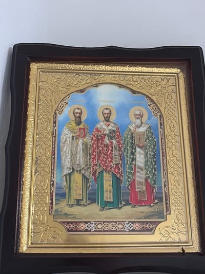 This image shows an Orthodox icon featuring three male saints standing side-by-side against a blue sky. The saints are depicted from the waist up, each with a golden halo. The saint on the left wears a white and gold robe and holds a red book. The middle saint is dressed in a red and gold robe, also holding a red book. The saint on the right wears a green and gold robe and holds a book with an ornate cover. The icon is framed by an ornate gold border with intricate geometric patterns. The entire icon is housed in a dark wooden frame. The image is an interior shot, with the icon hanging on a white wall. There is no visible indication of the city of Orhei, Moldova in this image.