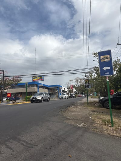 The image shows a street corner in San Rafael, Costa Rica (latitude: 9.937, longitude: -84.152). The street is paved and has a few cars driving on it. There are several businesses on the street, including a McDonald's, an am/pm gas station, and a sign advertising "SANGUICHOS" (sandwiches). The sky is partly cloudy, with a light blue color. A green pole with a sign reading "20 M" is in the foreground. There is a person walking near the McDonald's.