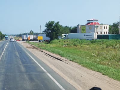 The image shows a road in Chernorech'e, Russia. The road is a two-lane asphalt road with a white line dividing the lanes. There are several trucks and cars on the road, as well as a building with a red roof. The building appears to be a business with signs that read "HEGELMANN GROUP" and "ЕВРО 24/7 Шиномонтаж". The surrounding area is mostly green grass and trees. The sky is blue and clear.