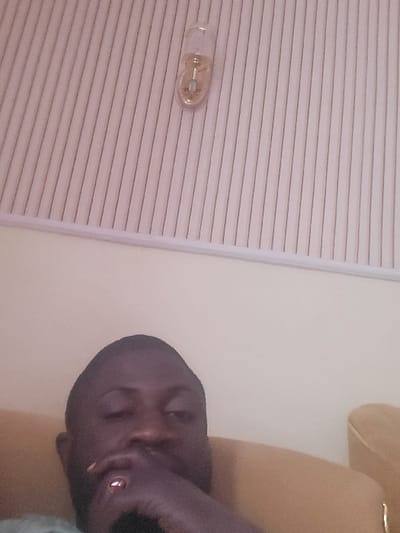 An indoor scene in Babawa, Nigeria, depicts a dark-skinned male subject, approximately mid-frame to the lower left, reclining on a tan or yellow upholstered chair or couch. The subject's left hand, adorned with a ring featuring a reddish stone on the ring finger, is positioned with fingers covering the mouth and chin area. His eyes appear partially closed. The background consists of a plain, light-colored wall (cream or light beige) below a white ceiling or upper wall section with vertical textured paneling. A single, clear capsule-shaped light fixture containing an internal bulb is mounted on the upper textured section. The overall lighting is dim and ambient.