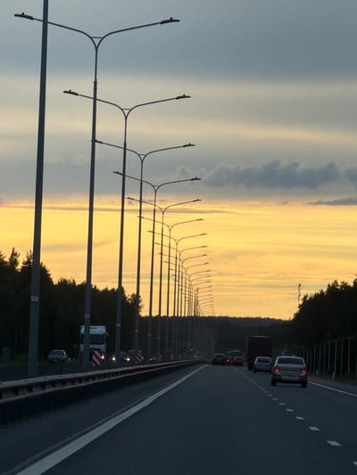 The image shows a highway at dusk, with a dramatic sunset casting a warm yellow and orange glow across the sky. The clouds are streaked and layered, creating a textured backdrop for the vibrant colors. The highway itself is dark asphalt, with white lane markings visible. A line of streetlights with a distinctive curved design runs along the left side of the road.
Several vehicles are visible on the highway, including cars and trucks. Some of the vehicles have their headlights on, suggesting the approaching night. The cars appear to be moving in the same direction, following the flow of traffic.
To the right of the highway, there is a dense line of trees, their dark silhouettes contrasting with the bright sky. The overall atmosphere is one of transition, as day gives way to night. The scene evokes a sense of travel and the vastness of the open road. There are no people clearly visible.
There is some faint text on small white markers attached to the street light poles, and on the back of some vehicles, but it is illegible. The image was taken in Brilliant, Russia.