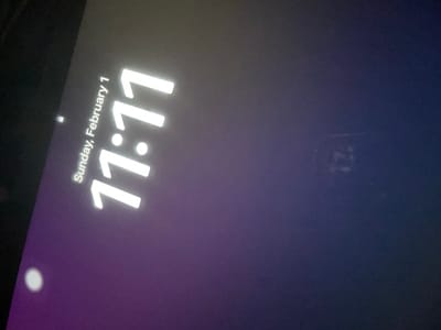 The image displays a digital screen, tilted, showing a lock screen interface. The screen exhibits a gradient background, transitioning from dark purple on the left to dark blue on the right. Prominently displayed in large white characters is the time "11:17". Above and to the left of the time, the date "Sunday, February 1" is shown in smaller white text. A small, white circular icon is present in the lower-left portion of the screen, with another similar, smaller indicator near the top-left. A faint, blurred, numerical artifact, possibly a reflection or ghosting resembling "12", is discernible in the upper-right quadrant. No visual elements within the image provide information regarding its specific location in Fegersheim, France.
