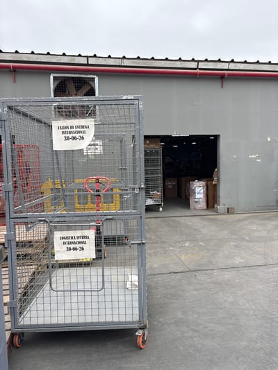 In Villa El Salvador, Peru, a gray warehouse has a large metal cage in the foreground. The cage is on wheels and has two signs attached to it. The top sign reads "FALLOS DE ENTREGA INTERNACIONAL 30-06-26". The bottom sign reads "LOGISTICA INVERSA INTERNACIONAL 30-06-26". Inside the cage, there are yellow and red pallet jacks and a red steering wheel. Beyond the cage, the warehouse opens into a loading bay where boxes and other items are stacked. A large exhaust fan is visible in a window above the entrance to the warehouse. The ground in front of the warehouse is concrete and shows tire marks.