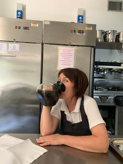 This media file shows a woman in what appears to be a commercial kitchen environment. She is wearing a white shirt and a dark apron, and is holding a glass coffee carafe up to her face, as if to smell or inspect its contents. The setting features stainless steel appliances, including what look like refrigerators with digital displays and control panels. There is text visible on the appliances and on paper labels attached to them. Some of the Italian text on the labels indicates "FRIGO" (refrigerator) and lists various food items such as "SALSA" (sauces), "SUGHI" (sauces/gravies), "ZUPPE" (soups), and "VERDURE COTTE" (cooked vegetables). Another label states, "E' VIETATO METTERE IN QUESTO FRIGO QUALSIASI PRODOTTO CHE NON SIA INERENTE AL SERVIZIO DELLE COLAZIONI (AD ESEMPIO BIBITE). GRAZIE PER LA COLLABORAZIONE," which translates to "It is forbidden to put in this refrigerator any product not related to the breakfast service (e.g. drinks). Thank you for your cooperation." In the background, a professional espresso machine is visible. The overall impression is a functional, possibly busy, food service establishment. The time of day and weather are not discernible from the image. The woman's expression is neutral.