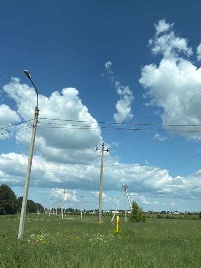 The image depicts an outdoor scene on a sunny day with a bright blue sky and scattered white clouds. In the foreground, a grassy field with wildflowers is visible. Several utility poles with power lines stretch across the frame. One prominent pole on the left has a street lamp attached to it. In the midground, more utility poles are spaced out across the field, leading towards a line of houses and trees in the distance. A yellow marker pole is also present in the midground. The overall environment suggests a rural or suburban landscape. The location is identified as Kamennoye, Russia.